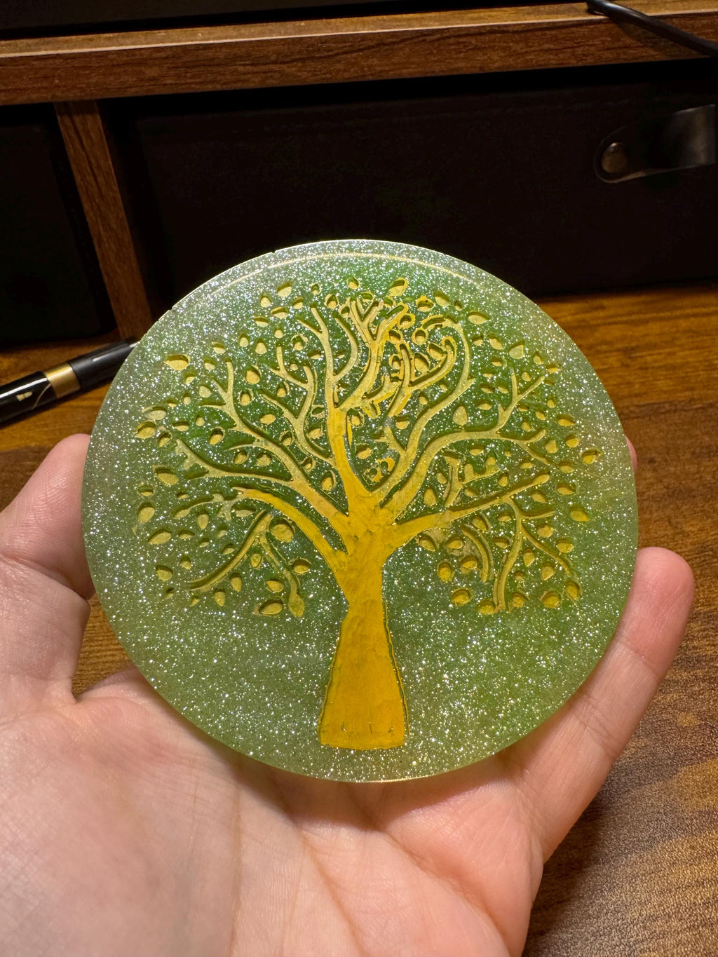 Hand holding a round green object with a yellow tree design on a resin surface